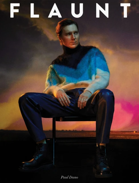 Paul Dano in a Flaunt magazine cover, seated on a chair wearing a multicolored sweater and leather pants, and black boots, in a studio.