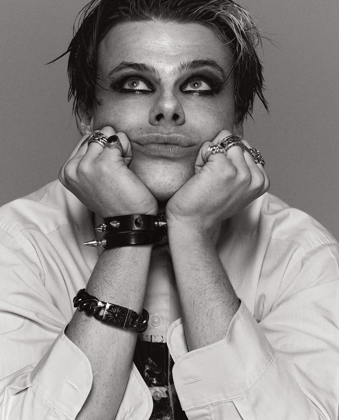 Black and white portrait of a young adult male with dark eye makeup, wearing a white button-down shirt and black bracelets, looking up.