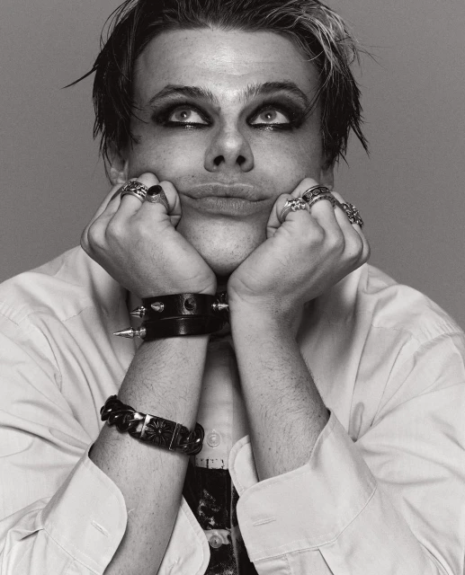 Black and white portrait of a young adult male with dark eye makeup, wearing a white button-down shirt and black bracelets, looking up.