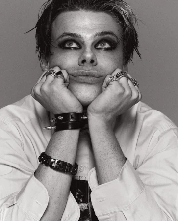 Black and white portrait of a young adult male with dark eye makeup, wearing a white button-down shirt and black bracelets, looking up.