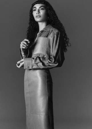 A woman poses in a monochromatic studio shot, wearing a satin top with large cuffs and a fitted leather skirt with visible seams.