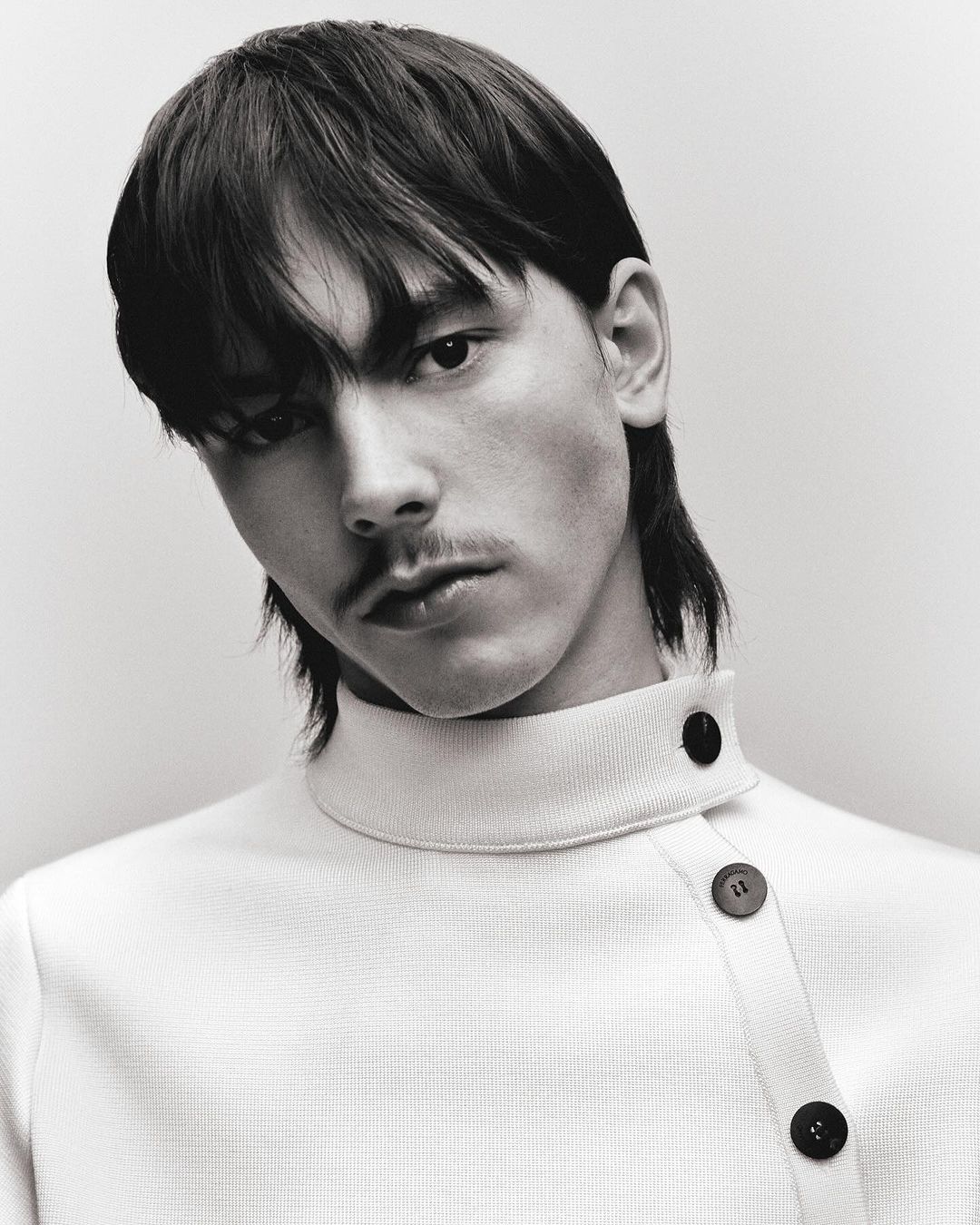 Portrait of a young adult male with dark hair and a mustache, wearing a cream-colored collared shirt with asymmetrical button detail.
