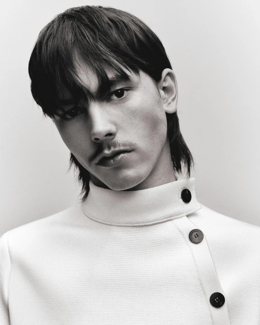 Portrait of a young adult male with dark hair and a mustache, wearing a cream-colored collared shirt with asymmetrical button detail.