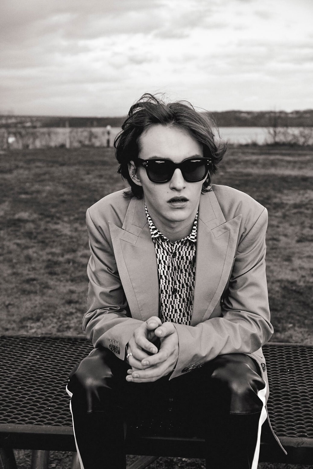 Monochrome portrait of a young adult man in sunglasses and a blazer, seated outdoors on a bench in front of a blurred background.