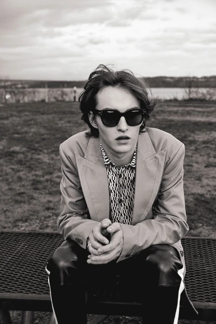 Monochrome portrait of a young adult man in sunglasses and a blazer, seated outdoors on a bench in front of a blurred background.