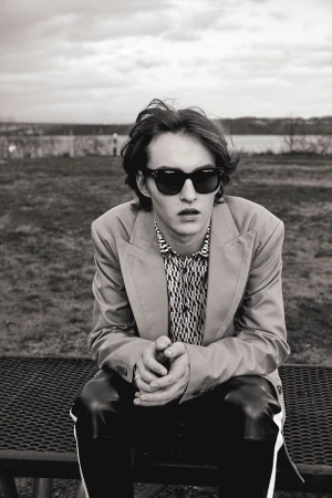 Monochrome portrait of a young adult man in sunglasses and a blazer, seated outdoors on a bench in front of a blurred background.