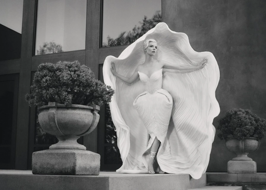 Elegant woman in a flowing white dress posing in front of a modern building with stone planters and greenery in a monochrome photograph.