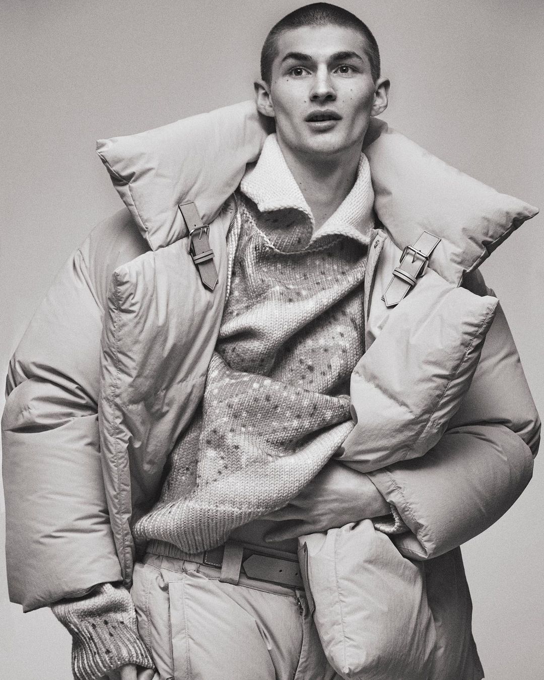 Monochrome portrait of a young male model in a puffy coat, textured sweater, and pale trousers, posing in a neutral studio backdrop.