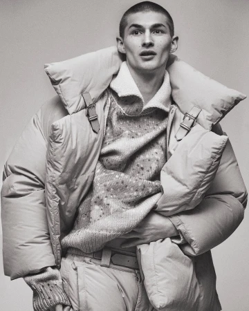 Monochrome portrait of a young male model in a puffy coat, textured sweater, and pale trousers, posing in a neutral studio backdrop.
