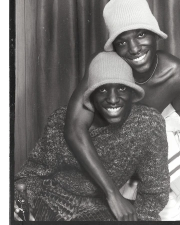 Black and white portrait of two Black adult males wearing hats and smiling, with one individual's arm around the other's shoulder.