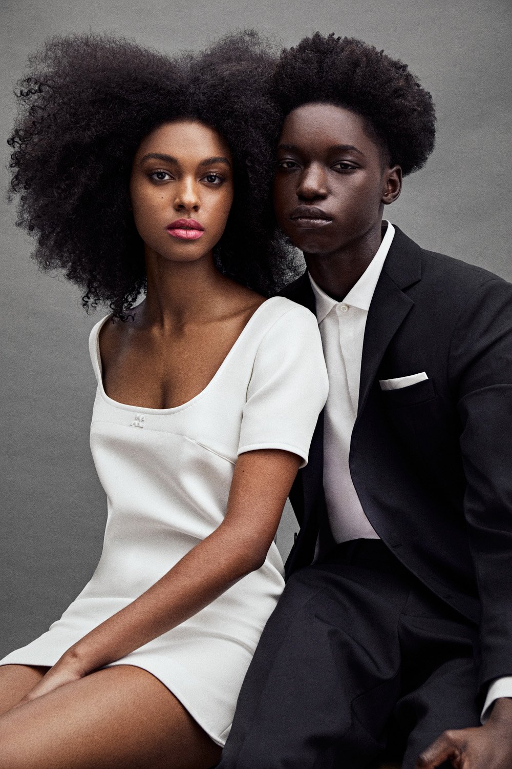 Portrait of a Black woman in a white dress next to a Black man in a black suit against a neutral background in a fashion editorial.