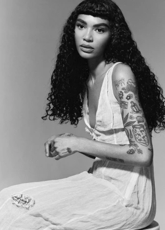 Black and white portrait of a woman with long curly hair and arm tattoos wearing a white dress against a grey studio backdrop.