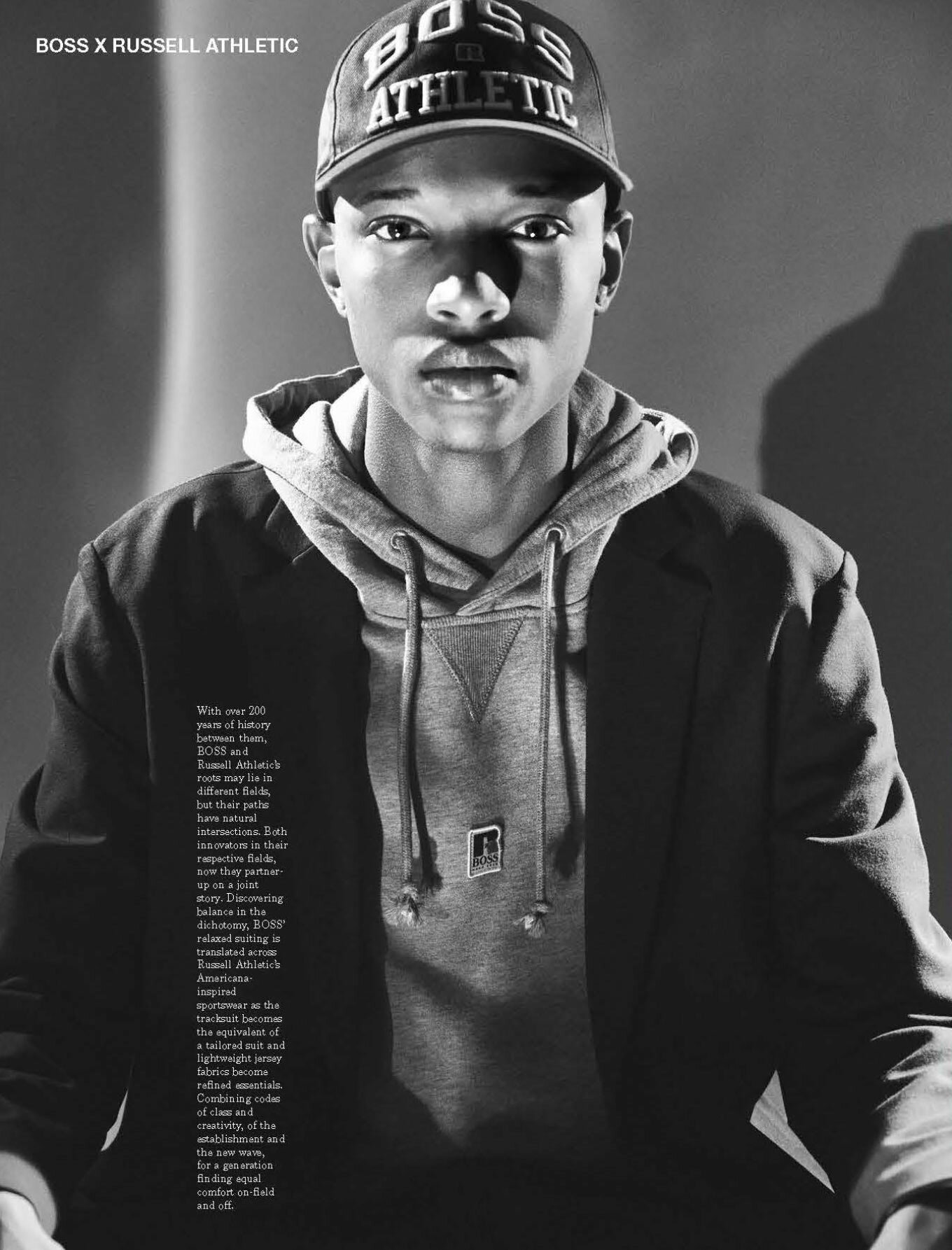 Black-and-white portrait of a young adult male wearing a cap, hoodie, and blazer. The model is posing and looking directly at the viewer.