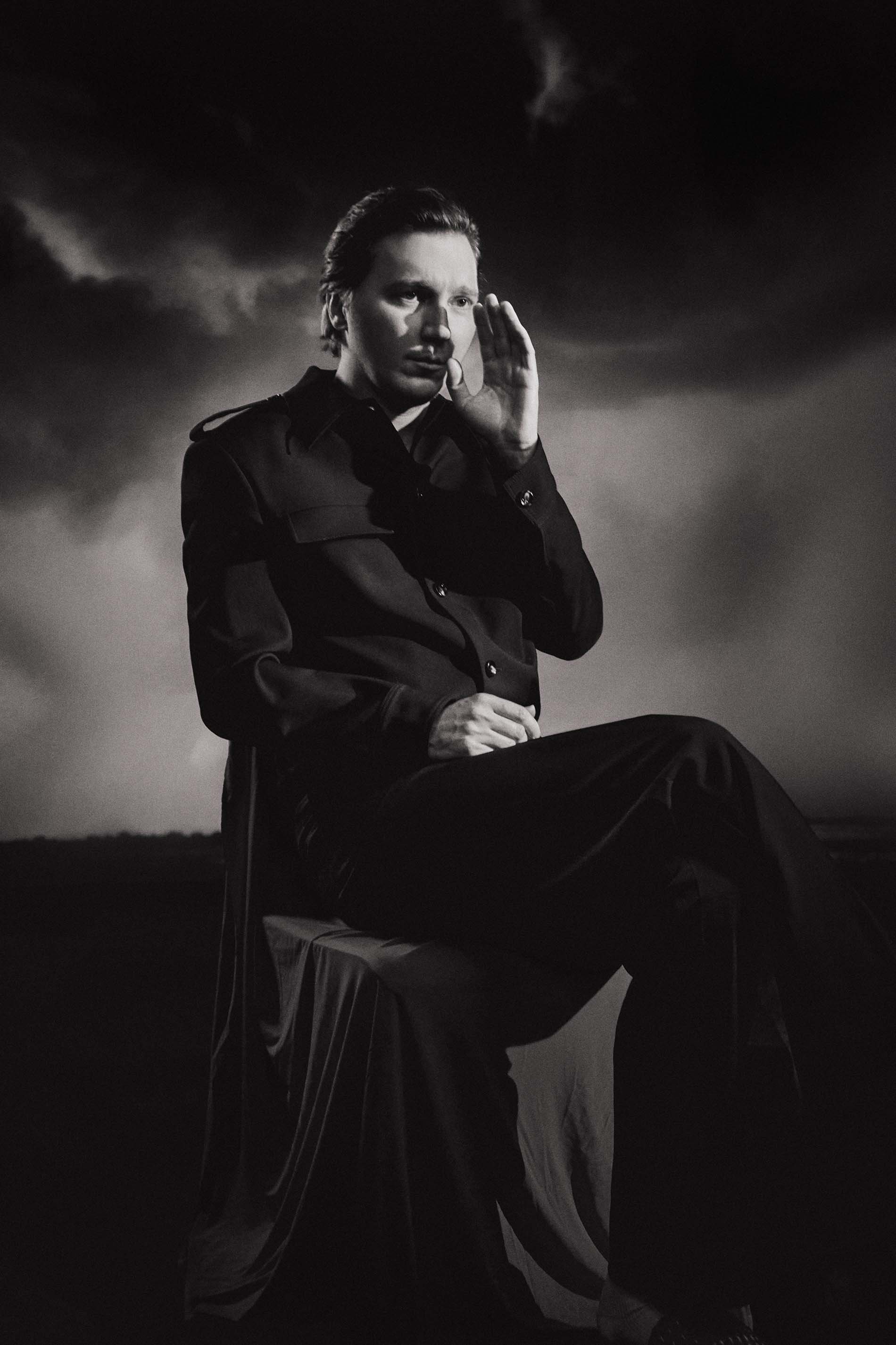 Monochromatic portrait of an adult male in dark clothing seated outdoors against a cloudy sky, hand raised to face.