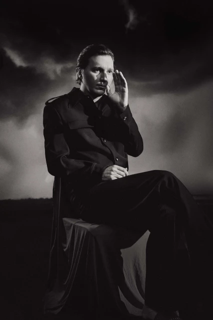 Monochromatic portrait of an adult male in dark clothing seated outdoors against a cloudy sky, hand raised to face.