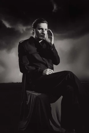Monochromatic portrait of an adult male in dark clothing seated outdoors against a cloudy sky, hand raised to face.