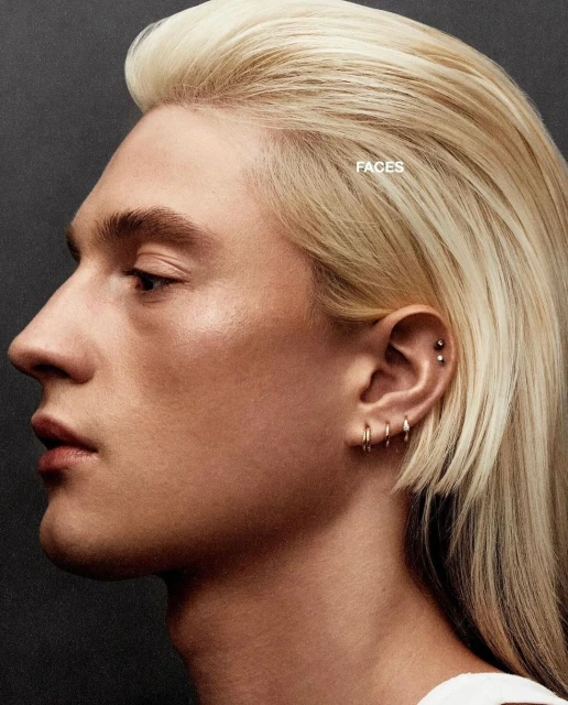 Profile portrait of a blonde adult male with multiple ear piercings, captured with studio lighting against a dark grey backdrop.