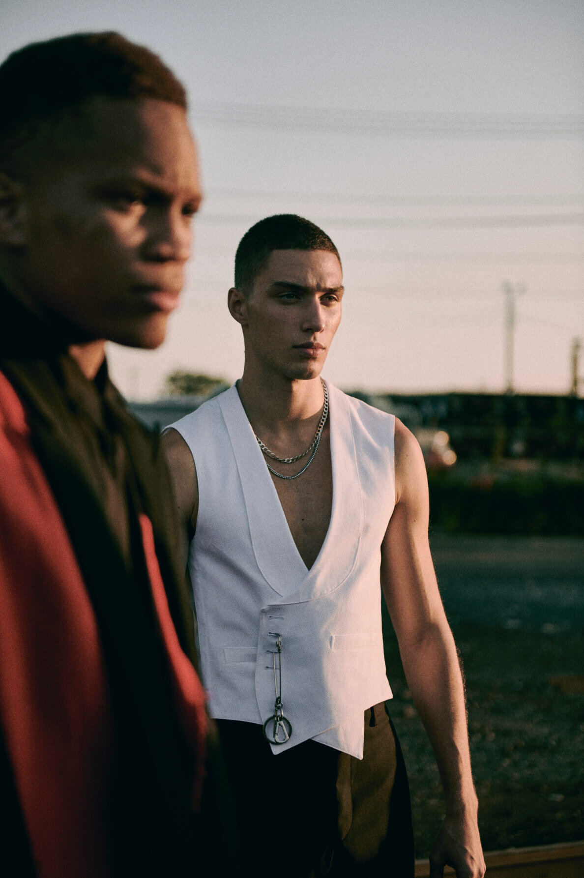 Two male models posing outdoors at sunset, one wearing a white vest and silver chain necklace with a second man blurred in the foreground.