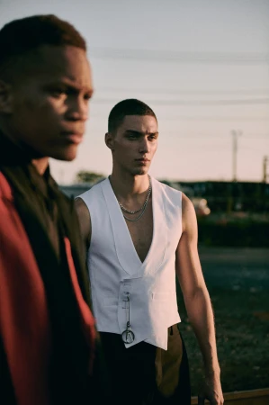 Two male models posing outdoors at sunset, one wearing a white vest and silver chain necklace with a second man blurred in the foreground.