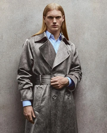 Portrait of a fair-skinned male fashion model with long blond hair, wearing a blue striped shirt and a trench coat against a textured wall.