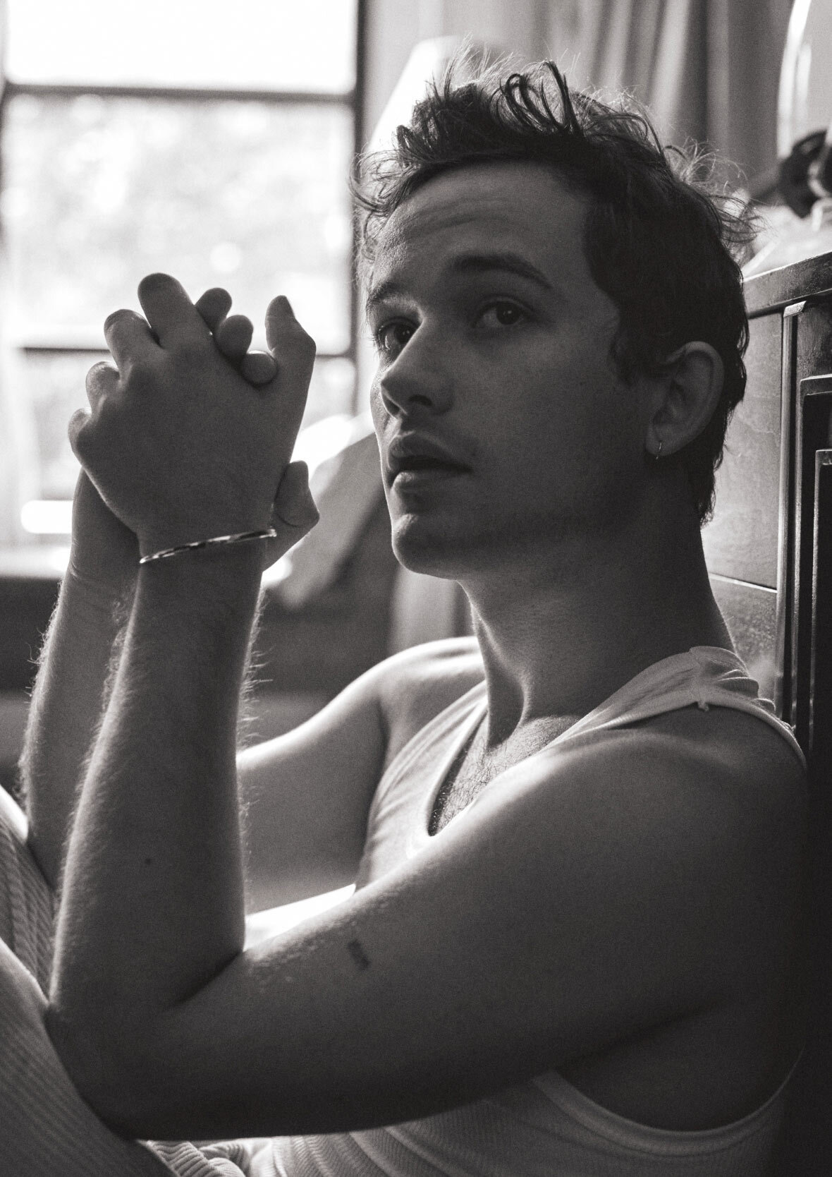 Black and white portrait of a fair-skinned adult male with tousled hair, wearing a tank top and bracelet, sitting indoors by a window.