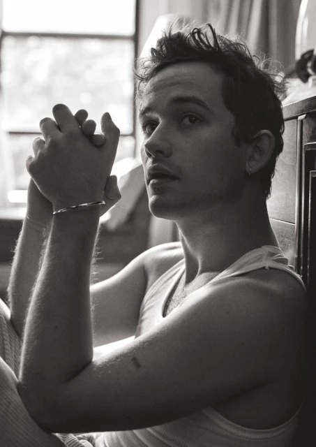 Black and white portrait of a fair-skinned adult male with tousled hair, wearing a tank top and bracelet, sitting indoors by a window.