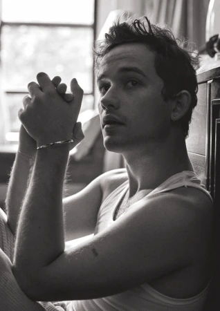Black and white portrait of a fair-skinned adult male with tousled hair, wearing a tank top and bracelet, sitting indoors by a window.