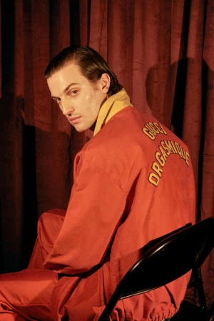A slender adult male sits on a black chair in a photo studio with a red curtain, wearing a red-orange tracksuit with Gucci text on the back.