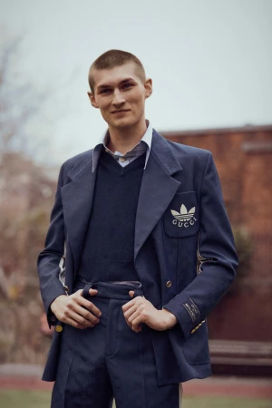 A man wearing a navy blazer and trousers with white branding details standing outdoors.