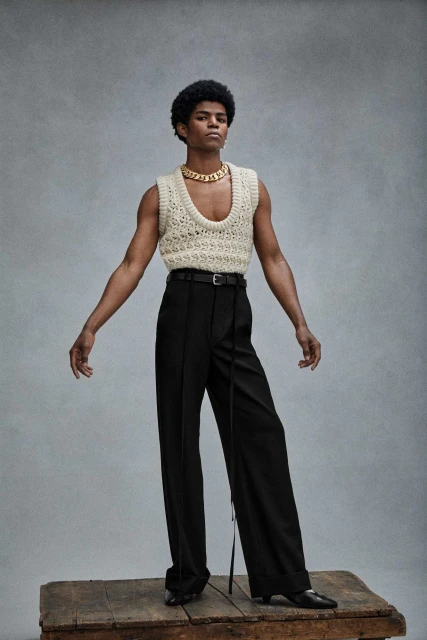 Studio shot of a Black male model wearing an ivory knit vest and black dress pants standing on a wood platform against a grey background.