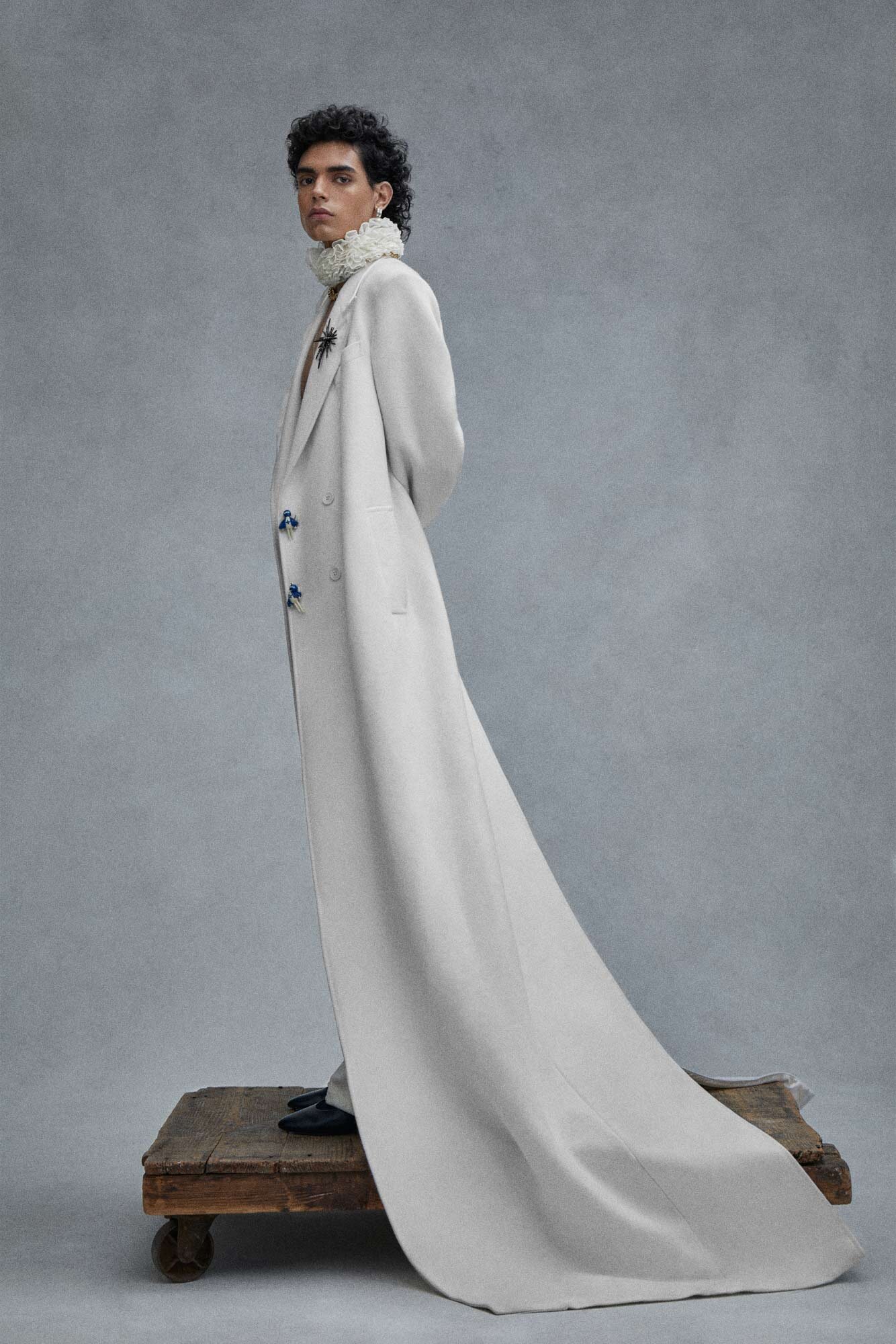 A fashion portrait showcases a person in a long cream coat with blue buttons, standing on a wooden platform against a grey backdrop.
