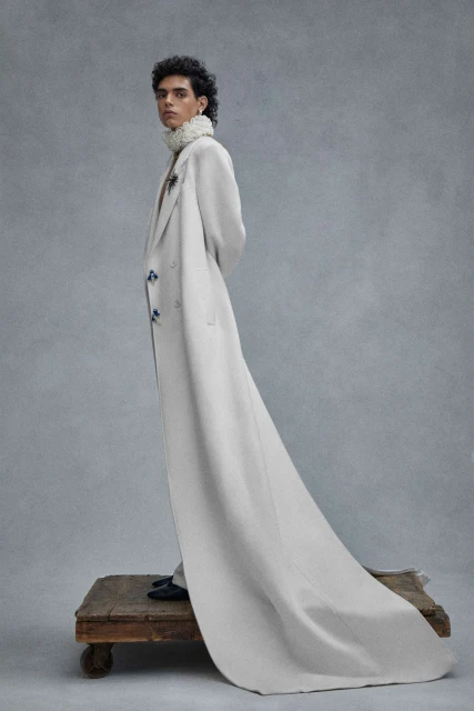 A fashion portrait showcases a person in a long cream coat with blue buttons, standing on a wooden platform against a grey backdrop.
