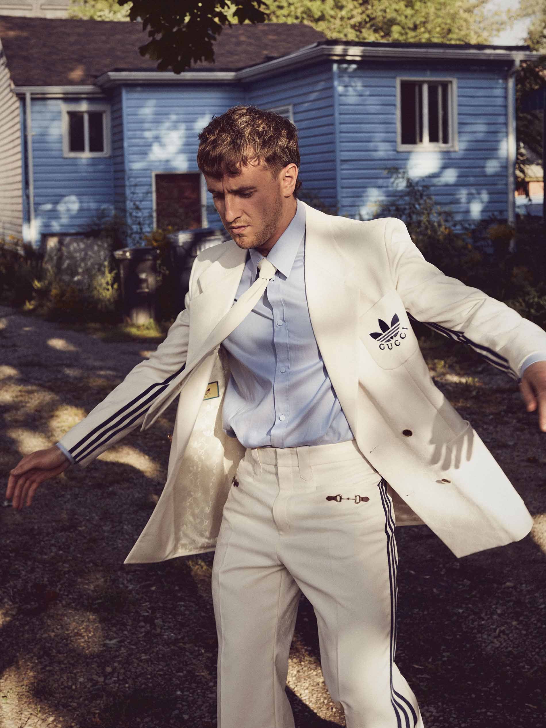 Man poses outdoors in a white Adidas x Gucci suit with black stripes and logos, wearing a blue shirt and standing outside of a house.