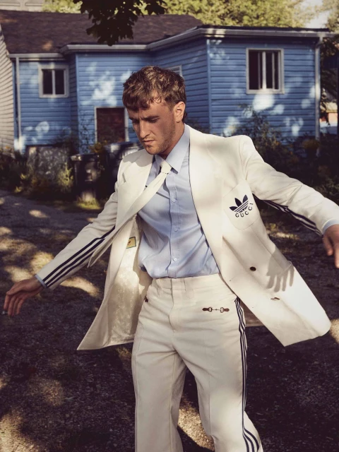 Man poses outdoors in a white Adidas x Gucci suit with black stripes and logos, wearing a blue shirt and standing outside of a house.