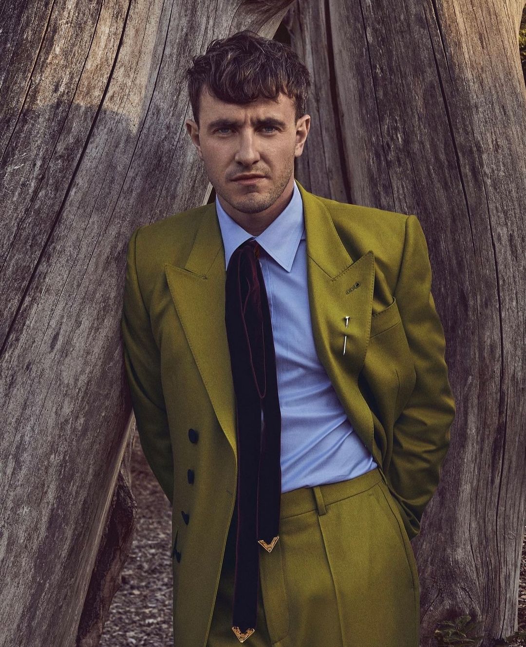 Portrait of a man in an olive green suit standing beside a weathered wooden structure, captured in soft, natural lighting.