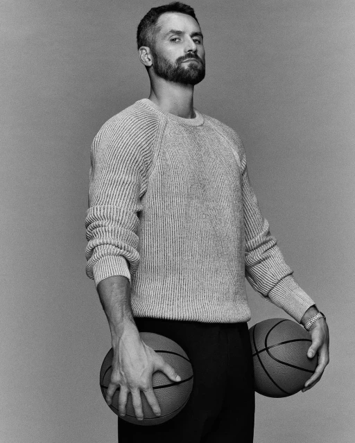Black and white portrait of a bearded adult male holding two basketballs, wearing a knit sweater and dark pants against a plain backdrop.