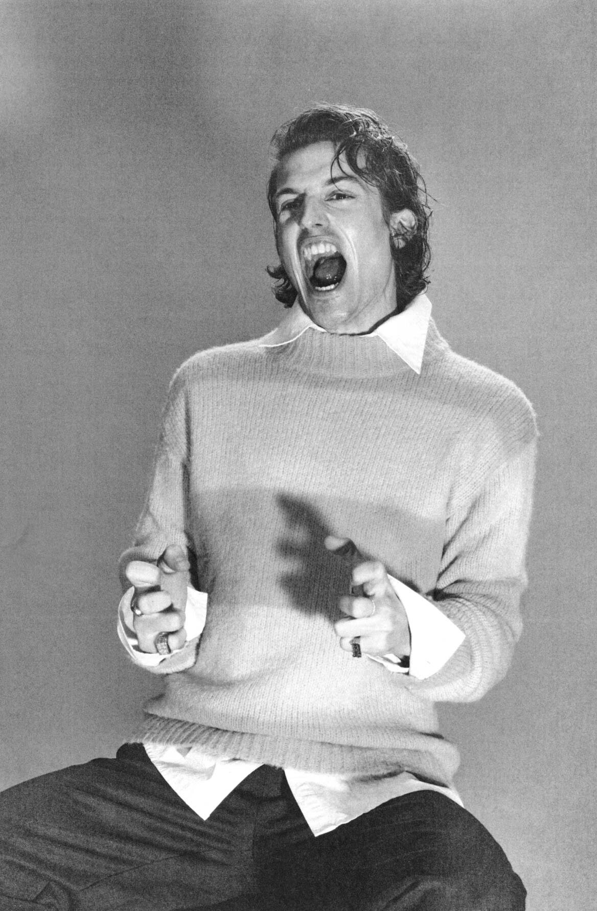 Black and white portrait of a young adult man with curly hair and open mouth, wearing a sweater and collared shirt against a gray backdrop.
