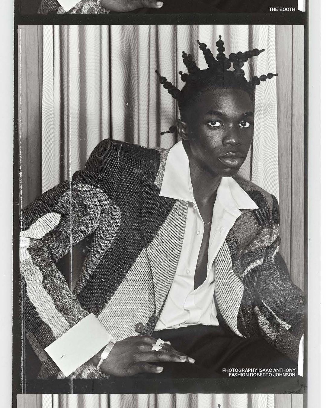 Monochromatic portrait of a dark-skinned man with unique hairstyle, wearing a blazer and unbuttoned shirt in a photo booth style.