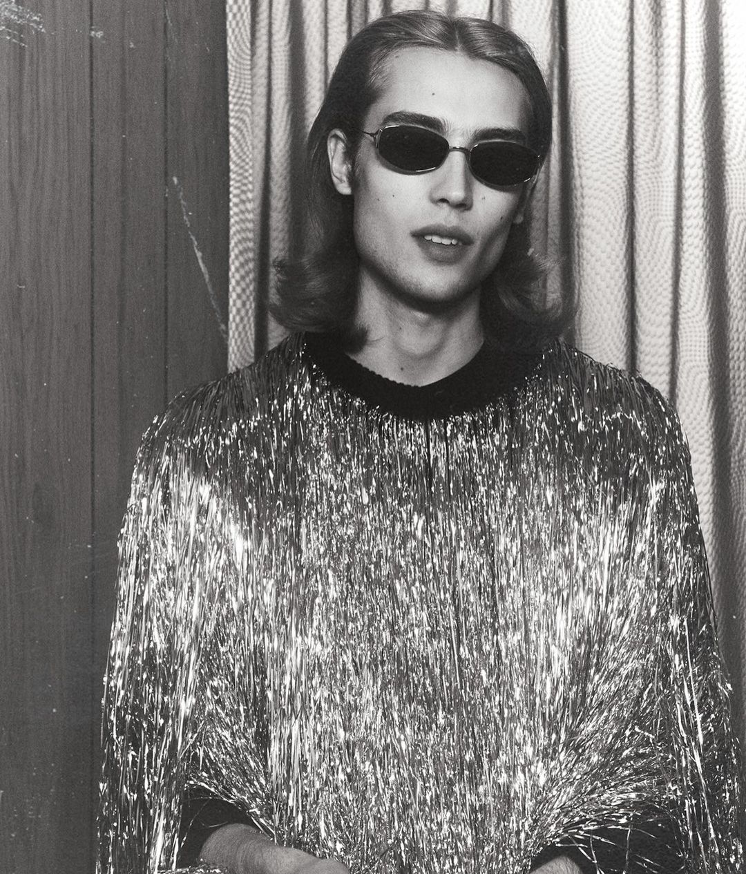 Monochrome portrait of an adult man wearing dark sunglasses and a silver tinsel shirt, set against a vertically striped background.
