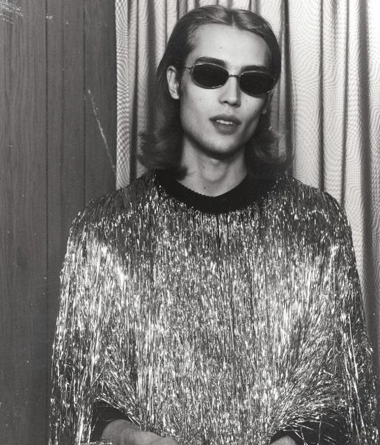 Monochrome portrait of an adult man wearing dark sunglasses and a silver tinsel shirt, set against a vertically striped background.