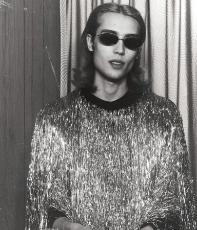 Monochrome portrait of an adult man wearing dark sunglasses and a silver tinsel shirt, set against a vertically striped background.