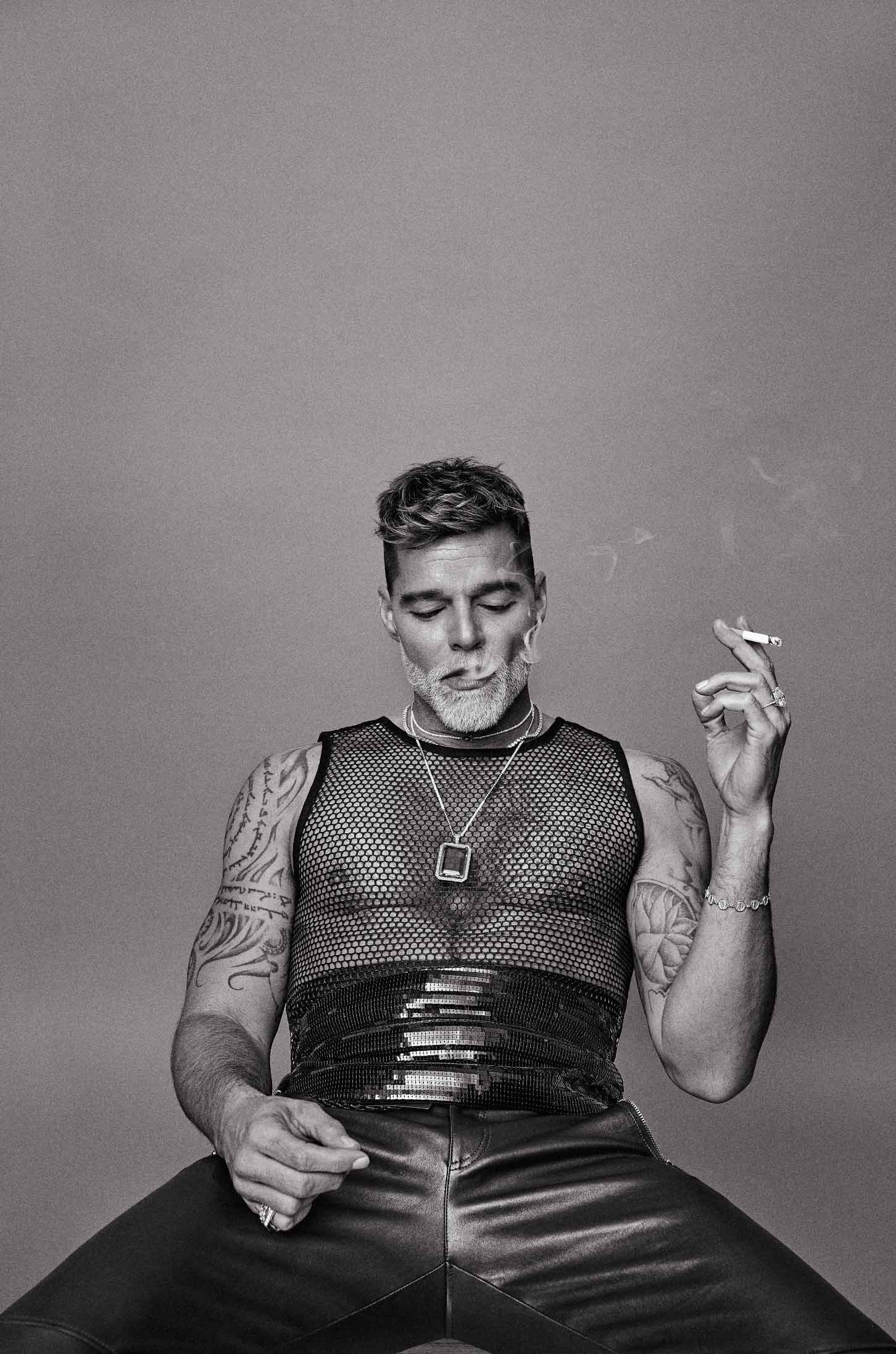 Monochrome portrait of an adult male seated, wearing a mesh top and leather pants, holding a lit cigarette, against a plain background.