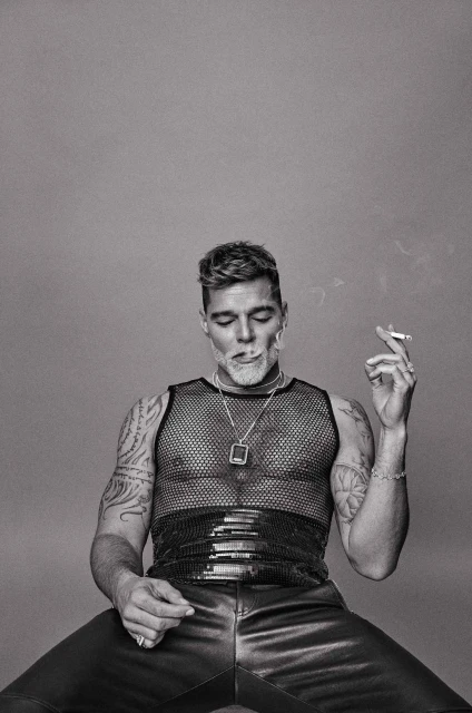 Monochrome portrait of an adult male seated, wearing a mesh top and leather pants, holding a lit cigarette, against a plain background.