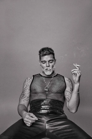 Monochrome portrait of an adult male seated, wearing a mesh top and leather pants, holding a lit cigarette, against a plain background.