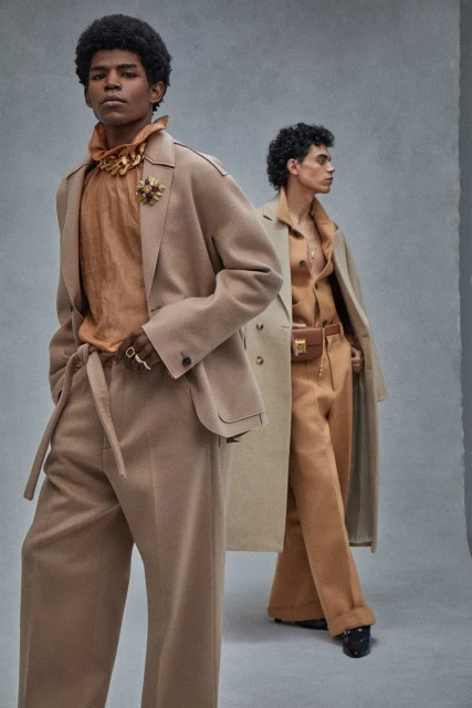 Two adult male models in beige colored suits posing indoors in front of a grey wall, promoting fashion and style in a studio setting.