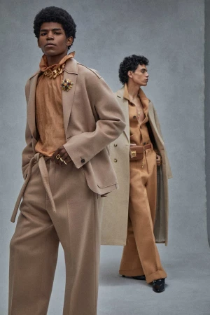 Two adult male models in beige colored suits posing indoors in front of a grey wall, promoting fashion and style in a studio setting.