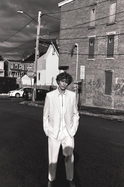 Monochrome portrait of a young man in a white suit standing on a city street with brick buildings and overcast sky in the background.