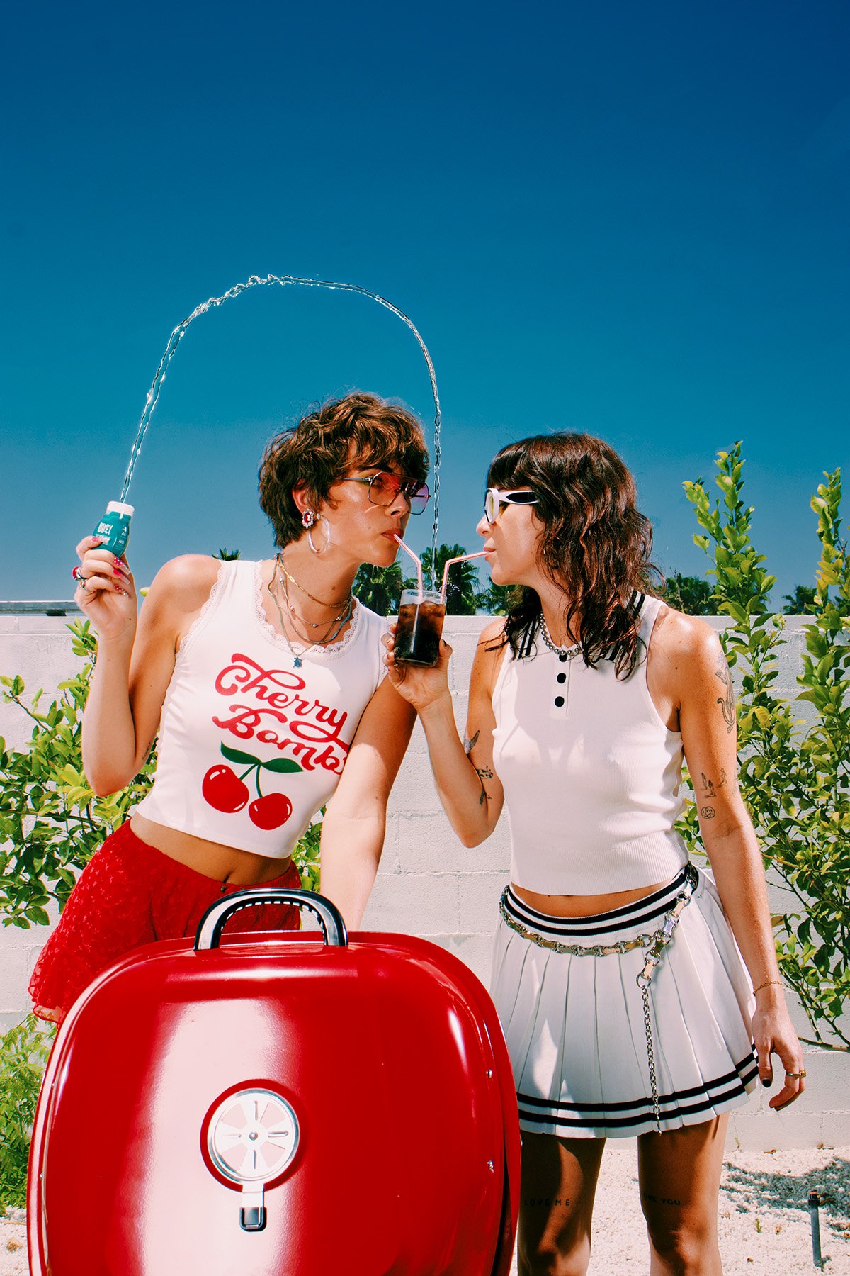 Two young women sharing a drink with straws outdoors next to a bright red grill under a clear blue sky