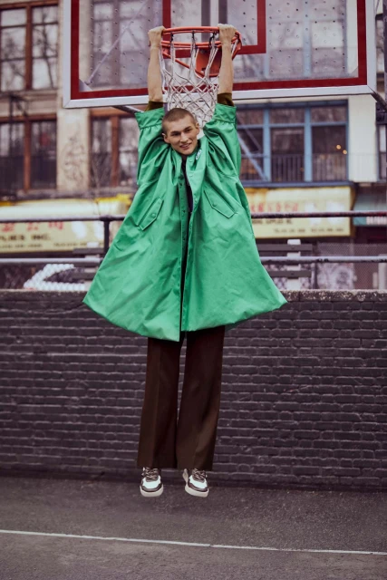 A person hanging from a basketball hoop on an outdoor court wearing a bright green coat and brown pants, with buildings in background.