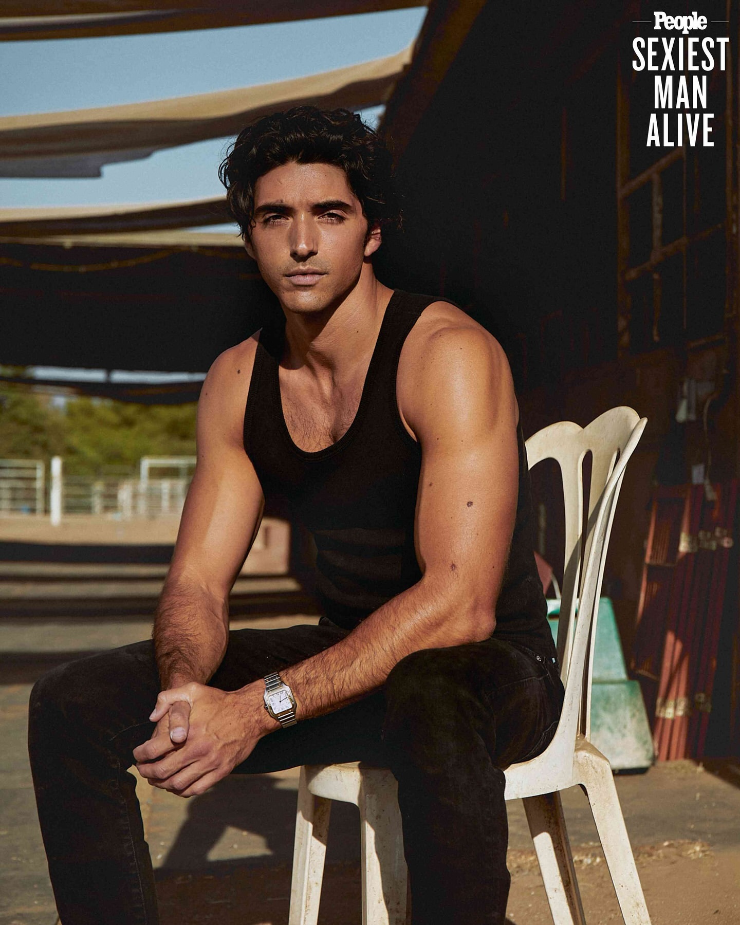 Portrait of a handsome man with dark hair in a black tank top sitting on a white chair outdoors, with a 'Sexiest Man Alive' banner.